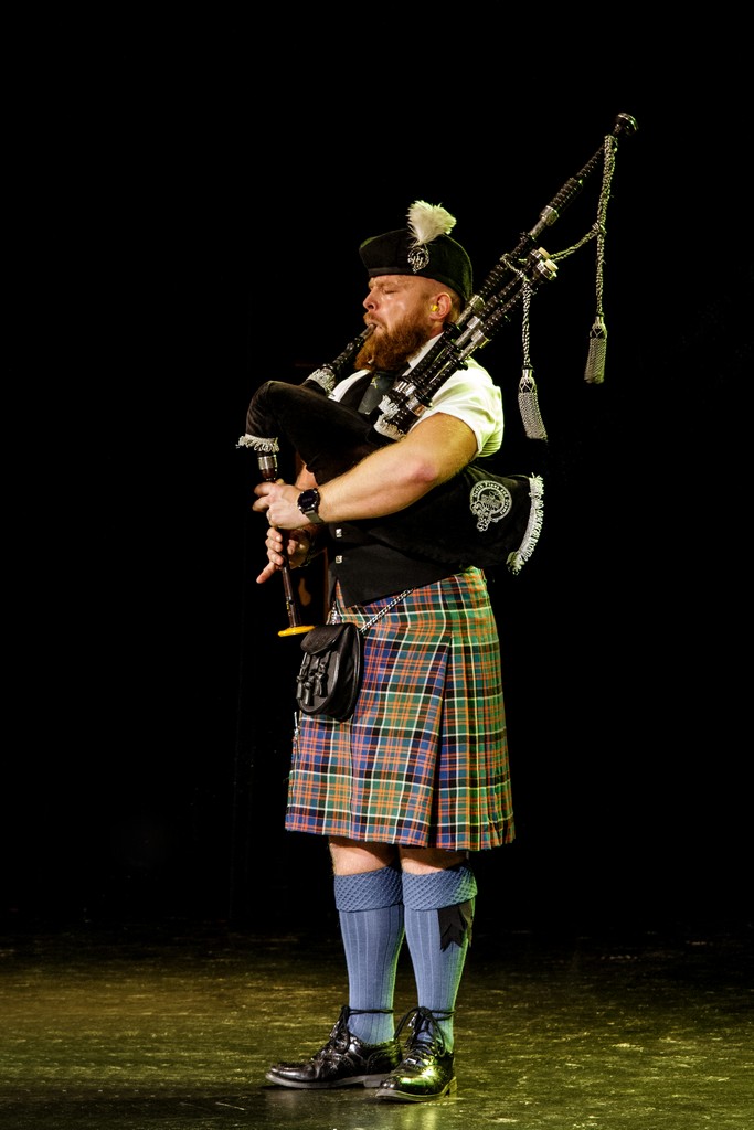 Bruce Korthout speelt solo doedelzak in wit overhemd, vest en kilt op een podium